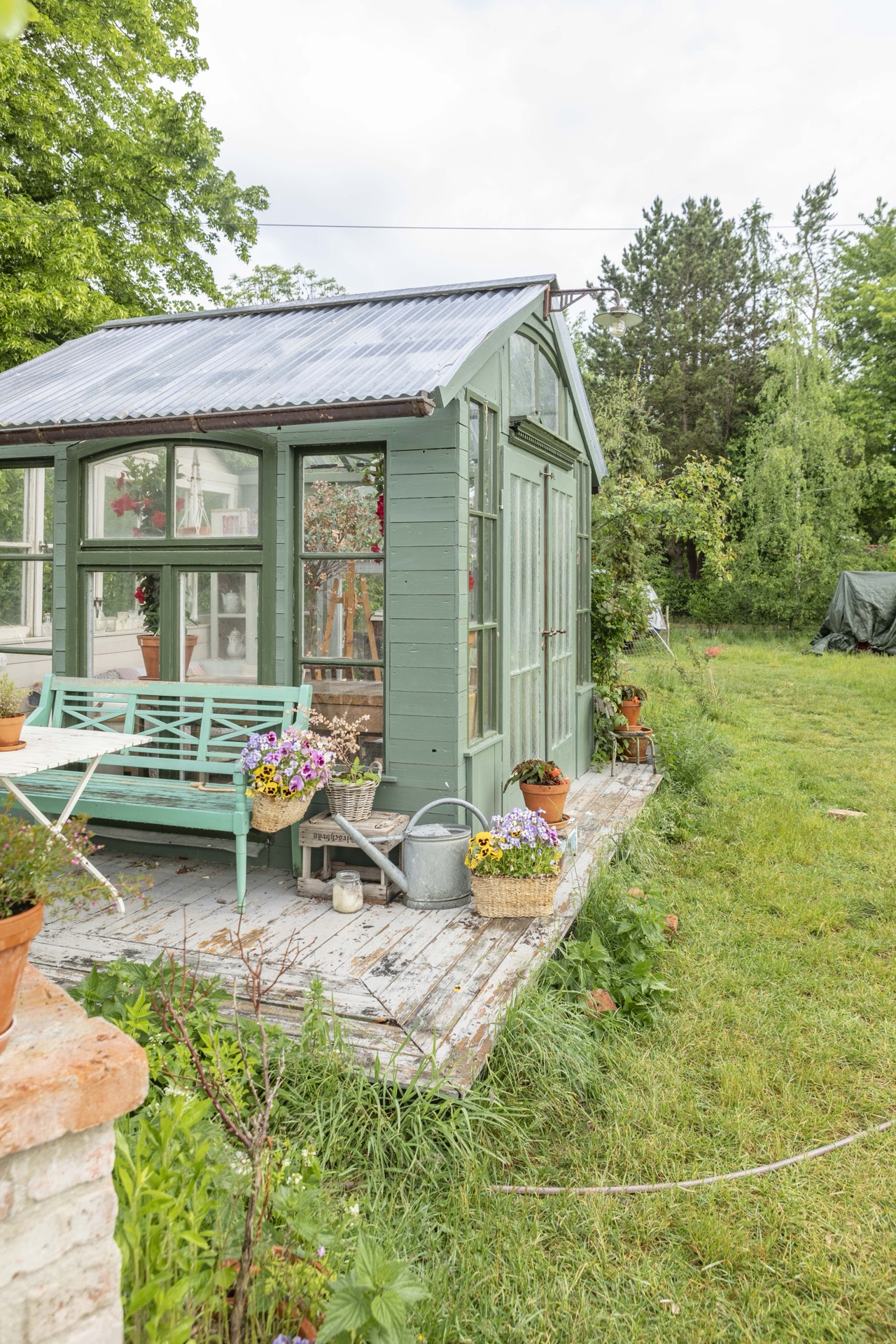 Salbeigrünes Gartenhaus mit Bank, Blumentöpfen und Gießkanne auf der Holzterrasse