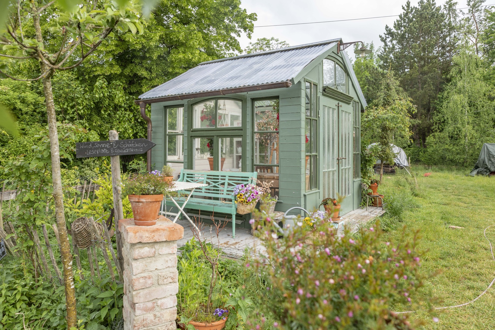 Salbeigrünes Gartenhaus mit gemauertem Pfeiler und blühenden Stauden im Vordergrund